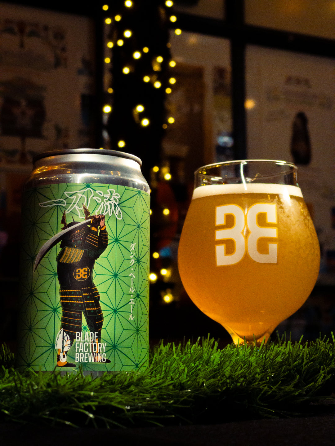 Can and glass of Blade Factory Brewing beer on grass with blurred lights in the background